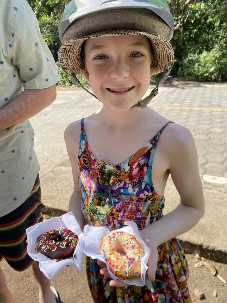 Girl in scooter helmet and colourful clothing holds two donuts with spinkles, one vanilla and one chocolate